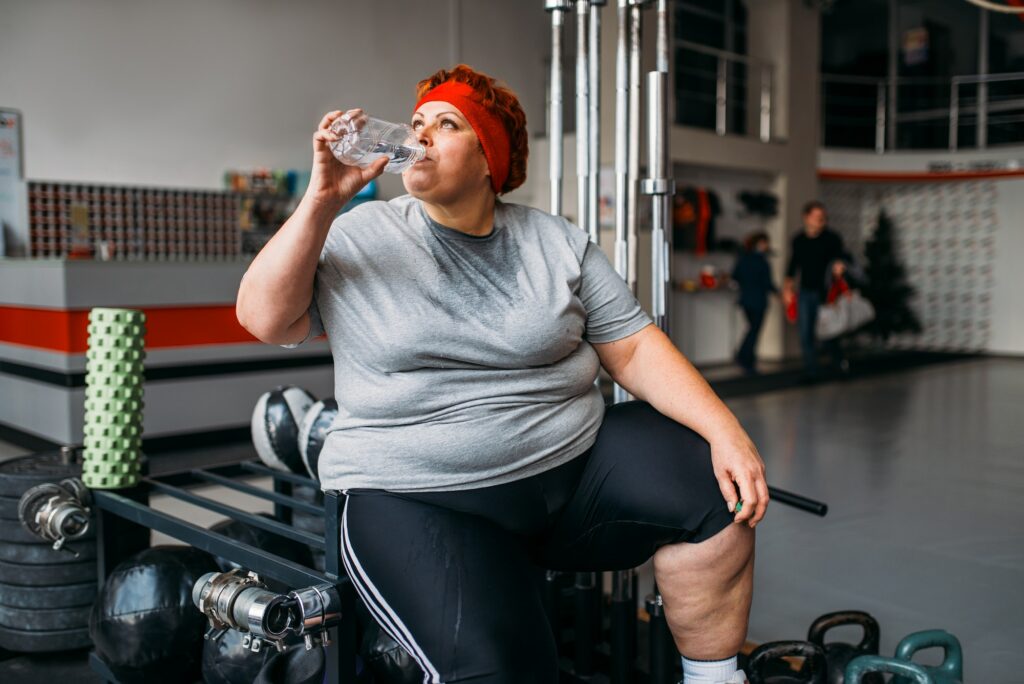 Fat woman drinks water after training in gym